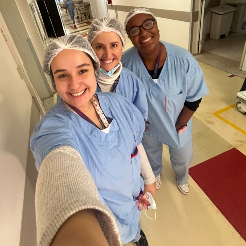 Mariana com colegas de equipe, de touca e pijama cirúrgico, no corredor do centro obstétrico